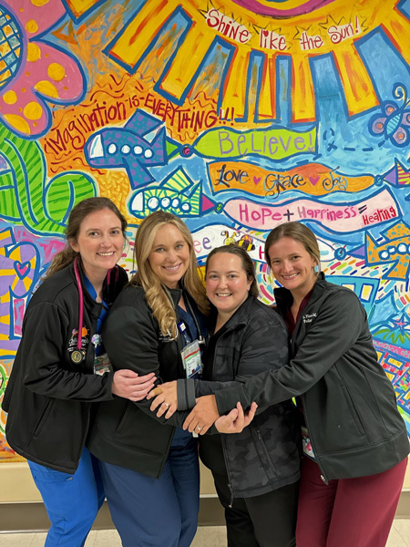 four residents, wearing scrubs and hugging, pose in front of a mural